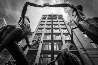 Two large statues of human figures are captured from a low angle, creating a dramatic perspective against a tall building with reflective windows. The statues are reaching out to each other, their arms forming an arch above. The composition is monochrome, highlighting the contrast between the gleaming sculptures and the architectural lines of the building.