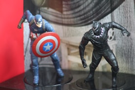 Two detailed action figures are positioned on a glossy black surface. The first figure, dressed in a blue costume with a star emblem, holds a circular shield with red, white, and blue colors. The second figure, clad in an all-black suit decorated with silver accents, stands poised for action. The background features a gray, textured pattern with a prominent red section on the left.
