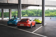 Three sports cars are parked in a covered outdoor area beside a modern building. The cars are brightly colored in blue, orange, and yellow. The area is surrounded by greenery, and there is branding related to Porsche on a barrier in the background.