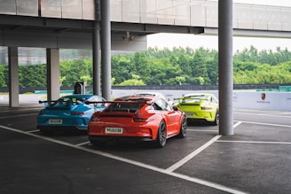 Three sports cars are parked in a covered outdoor area beside a modern building. The cars are brightly colored in blue, orange, and yellow. The area is surrounded by greenery, and there is branding related to Porsche on a barrier in the background.