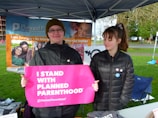 Two individuals are standing under a canopy with promotional banners in the background. One person is holding a bright pink sign that reads 'I Stand With Planned Parenthood.' Both individuals are wearing jackets, and the setting appears to be outdoors with green grass and a pathway visible.