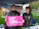 Two individuals are standing under a canopy with promotional banners in the background. One person is holding a bright pink sign that reads 'I Stand With Planned Parenthood.' Both individuals are wearing jackets, and the setting appears to be outdoors with green grass and a pathway visible.