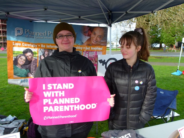 Two individuals are standing under a canopy with promotional banners in the background. One person is holding a bright pink sign that reads 'I Stand With Planned Parenthood.' Both individuals are wearing jackets, and the setting appears to be outdoors with green grass and a pathway visible.