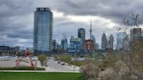 An urban skyline with modern high-rise buildings and the CN Tower in the background. A red abstract sculpture stands in the foreground near a green lawn, with a cloudy sky overhead. Bare trees with sparse leaves frame the scene, and a street lined with parked cars runs through the cityscape.