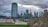 An urban skyline with modern high-rise buildings and the CN Tower in the background. A red abstract sculpture stands in the foreground near a green lawn, with a cloudy sky overhead. Bare trees with sparse leaves frame the scene, and a street lined with parked cars runs through the cityscape.