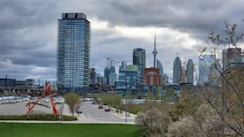 An urban skyline with modern high-rise buildings and the CN Tower in the background. A red abstract sculpture stands in the foreground near a green lawn, with a cloudy sky overhead. Bare trees with sparse leaves frame the scene, and a street lined with parked cars runs through the cityscape.