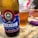 A close-up of a bottle of Eisenbahn American IPA beer placed on a countertop, alongside a glass filled with amber-colored beer. The bottle features a label with a vintage train emblem and text in Portuguese. The background includes a knife and some packaged food items.