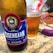 A close-up of a bottle of Eisenbahn American IPA beer placed on a countertop, alongside a glass filled with amber-colored beer. The bottle features a label with a vintage train emblem and text in Portuguese. The background includes a knife and some packaged food items.