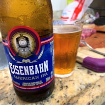 A close-up of a bottle of Eisenbahn American IPA beer placed on a countertop, alongside a glass filled with amber-colored beer. The bottle features a label with a vintage train emblem and text in Portuguese. The background includes a knife and some packaged food items.
