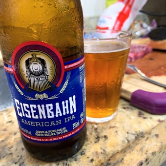 A close-up of a bottle of Eisenbahn American IPA beer placed on a countertop, alongside a glass filled with amber-colored beer. The bottle features a label with a vintage train emblem and text in Portuguese. The background includes a knife and some packaged food items.