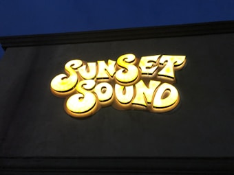 A neon sign displays the words 'Sunset Sound' in large, bold letters with a glowing yellow color against a dark background. The letters have a retro, whimsical style.
