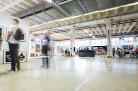 An art gallery interior features various artworks displayed on the walls and stands. Several people are seen interacting with the exhibits, with motion blur capturing their movements. The open, industrial-style space has large windows allowing natural light to brighten the room.