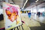 A colorful artwork on an easel depicts a profile of a person with abstract and vibrant strokes. The background consists of an indoor space with people walking in a large, open area featuring industrial design elements like exposed pipes and concrete flooring.