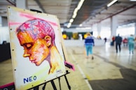 A colorful artwork on an easel depicts a profile of a person with abstract and vibrant strokes. The background consists of an indoor space with people walking in a large, open area featuring industrial design elements like exposed pipes and concrete flooring.