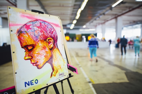 A colorful artwork on an easel depicts a profile of a person with abstract and vibrant strokes. The background consists of an indoor space with people walking in a large, open area featuring industrial design elements like exposed pipes and concrete flooring.