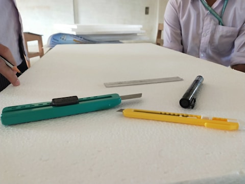 A flat surface with several tools and objects including a green utility knife, a yellow utility knife, a steel ruler, and a black marker. Two people are partially visible in the background, one in a white shirt standing on the left and another in a white shirt with a green lanyard sitting on the right. A stack of white boards or sheets is seen in the background.