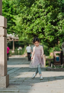 A young child wearing a light, sleeveless dress with floral patterns walks along a stone pathway in a park. The child is wearing leggings and white shoes. The background is lush with green foliage, and there are a couple of people walking further along the path, partially visible.