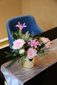 A floral arrangement with pink roses and lilies in a gold vase, adorned with green foliage, is placed on a table covered in a light-colored cloth. Behind the table, there is a blue cushioned chair.
