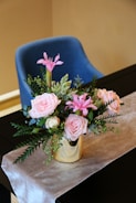 A floral arrangement with pink roses and lilies in a gold vase, adorned with green foliage, is placed on a table covered in a light-colored cloth. Behind the table, there is a blue cushioned chair.