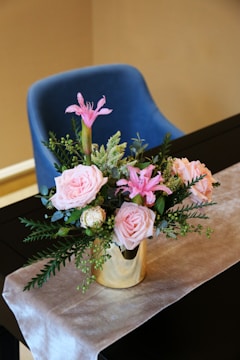 A floral arrangement with pink roses and lilies in a gold vase, adorned with green foliage, is placed on a table covered in a light-colored cloth. Behind the table, there is a blue cushioned chair.