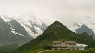 A serene alpine landscape featuring a quaint building nestled at the base of lush green hills. Snow-capped mountains tower impressively in the background under a cloudy gray sky.