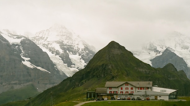 A serene alpine landscape featuring a quaint building nestled at the base of lush green hills. Snow-capped mountains tower impressively in the background under a cloudy gray sky.