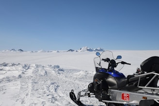 A snow-covered landscape with a snowmobile in the foreground, facing towards distant mountains. The sky is clear and blue, creating a crisp, tranquil scene.