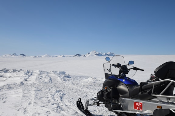 A snow-covered landscape with a snowmobile in the foreground, facing towards distant mountains. The sky is clear and blue, creating a crisp, tranquil scene.
