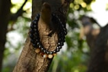 A string of beads is wrapped around the branch of a tree. The beads are predominantly dark with a glossy finish and some translucent ones in a mix of earth tones. The background consists of blurred greenery and dappled sunlight creating a serene atmosphere.