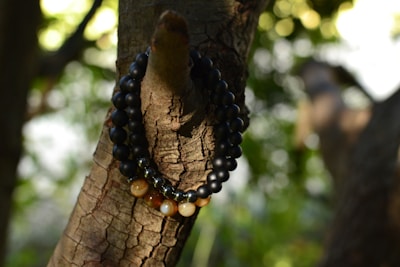 A string of beads is wrapped around the branch of a tree. The beads are predominantly dark with a glossy finish and some translucent ones in a mix of earth tones. The background consists of blurred greenery and dappled sunlight creating a serene atmosphere.