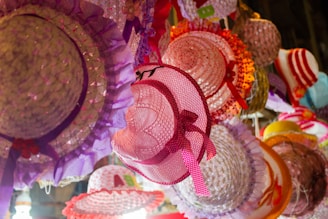 A collection of colorful hats hangs together. The hats feature intricate designs and bright colors, with some adorned with ribbons and decorative elements. The array includes shades of pink, purple, orange, and red, creating a vibrant and festive scene.