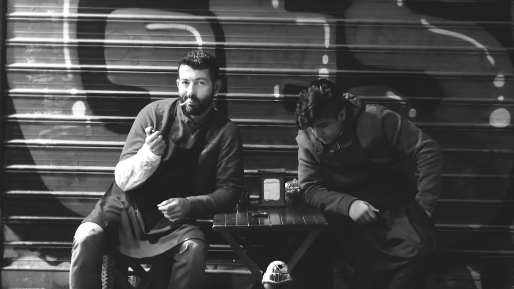 grayscale photo of two men sitting beside roller shutter