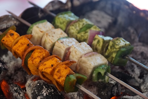 Three skewers of colorful vegetables and paneer are grilling over hot charcoal. The skewers include orange and green bell peppers, white paneer cubes, and bits of onion. The vivid colors of the vegetables contrast with the dark, ashy coals that have orange embers glowing underneath.