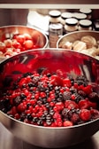 A large stainless steel bowl filled with a mixture of vibrant red and dark berries, including strawberries, raspberries, blackberries, and blackcurrants. In the background, two more bowls, one with cherries and another with what appears to be peeled or shelled nuts. Spice jars are partially visible.