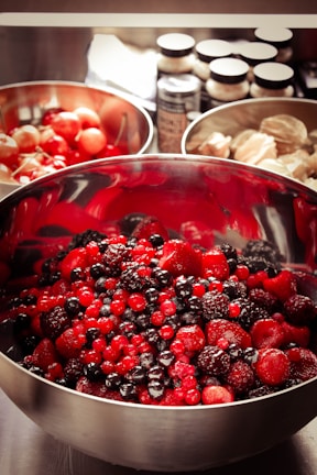 A large stainless steel bowl filled with a mixture of vibrant red and dark berries, including strawberries, raspberries, blackberries, and blackcurrants. In the background, two more bowls, one with cherries and another with what appears to be peeled or shelled nuts. Spice jars are partially visible.