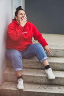 A person wearing a red hoodie and blue jeans is sitting on concrete steps, laughing. Their hair is tied up in a bun, and they are holding one hand near their mouth while their other hand rests on a step. They are wearing white sneakers and black socks.