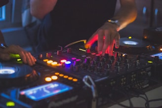 A DJ is mixing music on a professional DJ controller with multiple knobs and buttons. The scene is dimly lit, creating a vibrant and energetic atmosphere with colorful lights illuminating the equipment.