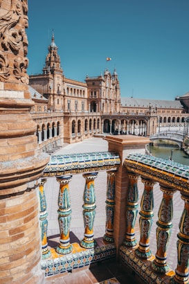 A grand architectural plaza with intricate ceramic tile work and ornate balustrades in the foreground, showcasing a historic building adorned with arches and towers. The scene is set under a clear blue sky, with a flag waving atop one tower and a glimpse of a water feature.
