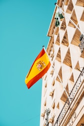 A Spanish flag flutters against a clear blue sky, mounted on an ornate historical building with classical architectural features. The facade is adorned with decorative details and geometric patterns.