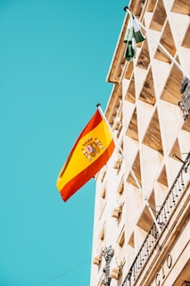 A Spanish flag flutters against a clear blue sky, mounted on an ornate historical building with classical architectural features. The facade is adorned with decorative details and geometric patterns.