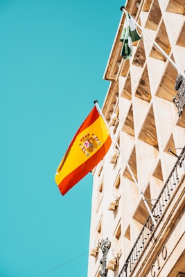 A Spanish flag flutters against a clear blue sky, mounted on an ornate historical building with classical architectural features. The facade is adorned with decorative details and geometric patterns.