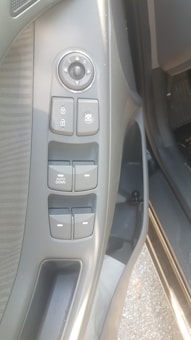 The image features the door panel of a car interior, showing multiple control buttons. The controls include a circular side mirror adjustment knob labeled with 'L' and 'R' for left and right, buttons for window lock and window adjustment, and an 'auto down' button specifically for automatic window lowering. The panel is mainly gray with a textured area near the top.