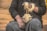 A person dressed in dark clothing is holding a spindle and wool. The wool is wrapped around the spindle as the person spins it into yarn. The scene has an earthy background with warm tones, and the person's hands are prominently featured engaging in the traditional craft.