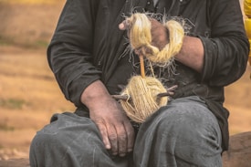 A person dressed in dark clothing is holding a spindle and wool. The wool is wrapped around the spindle as the person spins it into yarn. The scene has an earthy background with warm tones, and the person's hands are prominently featured engaging in the traditional craft.