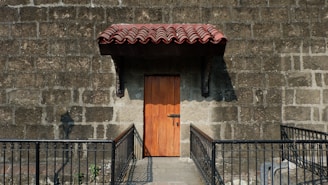 A wooden door with a metal lock set in a stone wall with a prominent red tiled awning above it. The area is bordered by a metal railing, and sunlight casts shadows on the stone wall.