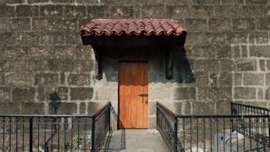 A wooden door with a metal lock set in a stone wall with a prominent red tiled awning above it. The area is bordered by a metal railing, and sunlight casts shadows on the stone wall.