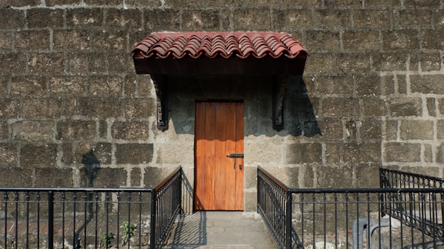 A wooden door with a metal lock set in a stone wall with a prominent red tiled awning above it. The area is bordered by a metal railing, and sunlight casts shadows on the stone wall.