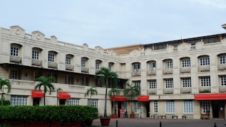 A multi-story building with an elegant architectural style featuring numerous windows and small balconies. The facade is painted in a light cream color, and red awnings complement the look. Tall green palm trees are planted in front of the building, and neatly trimmed hedges line the base. The courtyard area is paved, and people are casually interacting near the entrance.