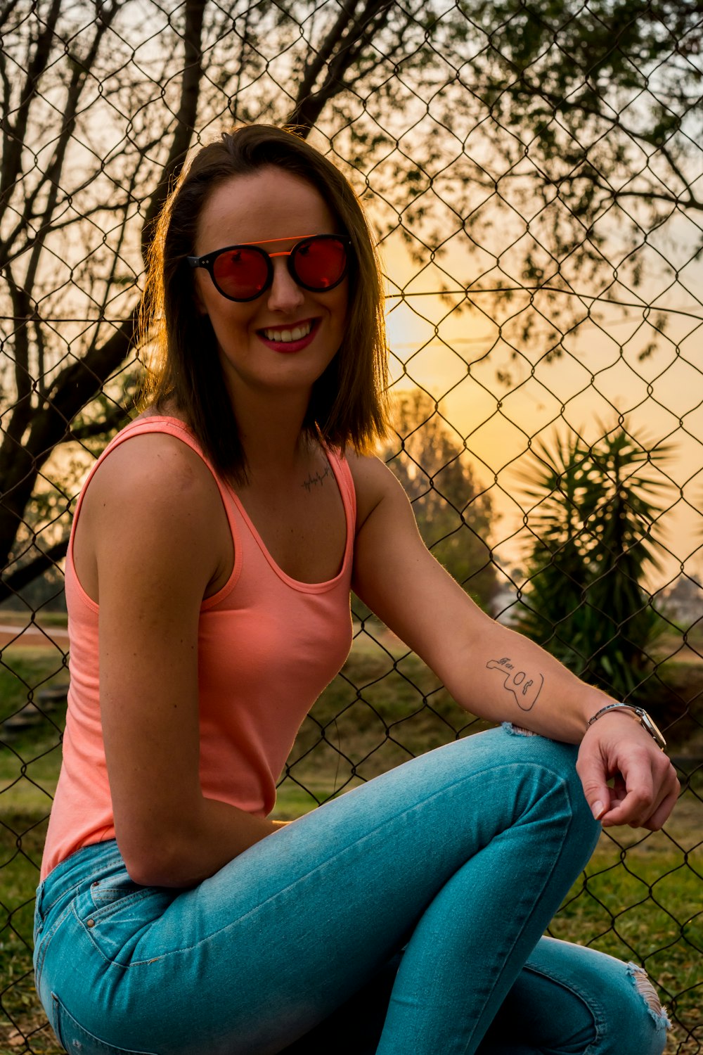 smiling woman in peach tank top and blue skinny denim jeans sitting near wire mesh fence