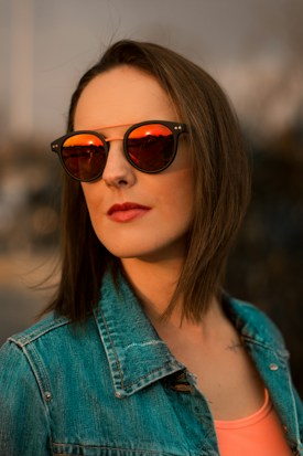 A person wearing reflective sunglasses that show a vibrant orange hue, along with a denim jacket and a light top. The warm lighting casts a dramatic shadow.